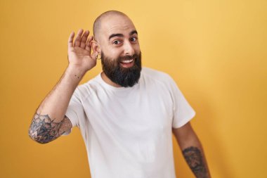 Sarı arka planda dikilen sakallı ve dövmeli genç İspanyol bir adam. Kulağı kulağında bir söylenti ya da dedikoduyu dinliyor. Sağırlık kavramı. 