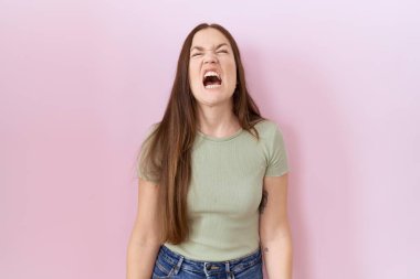 Beautiful brunette woman standing over pink background angry and mad screaming frustrated and furious, shouting with anger. rage and aggressive concept. 