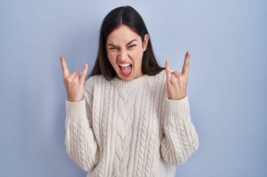 Young brunette woman standing over blue background shouting with crazy expression doing rock symbol with hands up. music star. heavy music concept. 