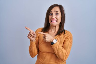 Orta yaşlı esmer kadın turuncu kazak giymiş endişeli ve gergin bir şekilde iki elini de işaret ediyor. Endişeli ve şaşırmış bir ifade. 