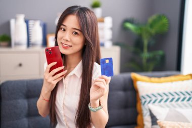 Young chinese woman using smartphone and credit card sitting on sofa at home