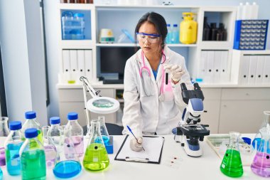 Young chinese woman wearing scientist uniform holding blood test tube writing on document at laboratory