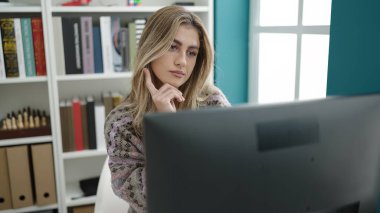 Young blonde woman student using computer studying with doubt expression at library university