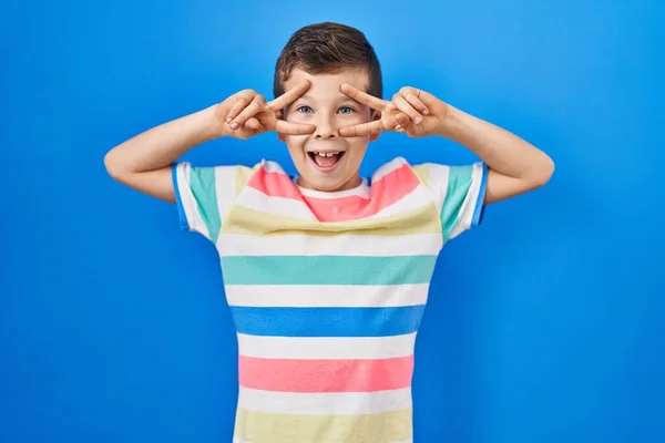青い背景の上に立つ若い白人の子供顔の上に指で平和のシンボルをやって 勝利を示す明るい笑顔 — ストック写真