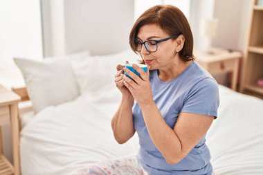 Orta yaşlı bir kadın yatak odasında bir fincan kahve içiyor.