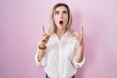 Young beautiful woman standing over pink background amazed and surprised looking up and pointing with fingers and raised arms. 