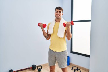 Young hispanic man smiling confident using dumbbells training at sport center