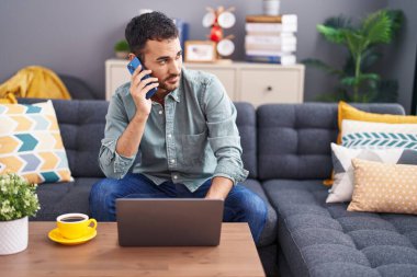 Young hispanic man talking on smartphone using laptop at home
