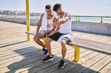Two hispanic men sporty couple smiling confident looking watch sitting on bench at seaside