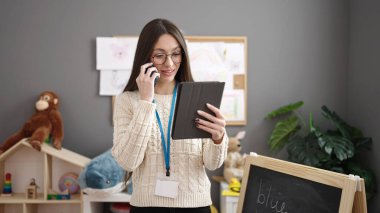 Young beautiful hispanic woman preschool teacher talking on smartphone using touchpad at kindergarten