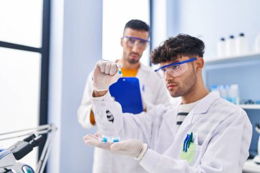 Elinde haplar tutan iki bilim adamı laboratuarda çalışan bir panoya yazı yazıyorlar.