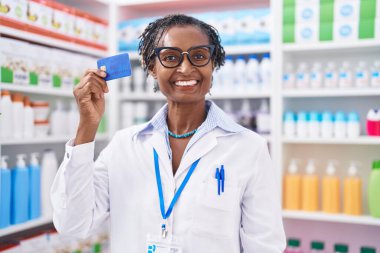 Eczanede çalışan Afrikalı bir kadın elinde kredi kartıyla mutlu ve pozitif görünüyor. Dişlerini gösteren bir gülümsemeyle ayakta duruyor. 