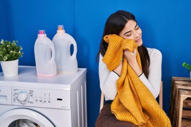 Young beautiful hispanic woman smiling confident touching softener clothes with face at laundry room