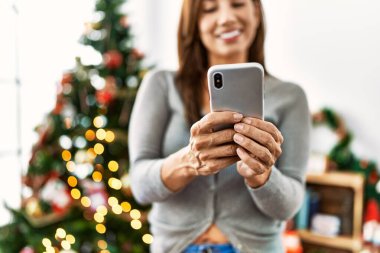 Akıllı telefon kullanan genç Latin kadın Noel ağacının yanında duruyor.