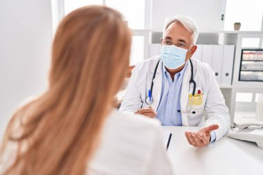 Orta yaşlı erkek ve kadın doktor ve hasta tıbbi maske takıyor klinikte danışmanlık yapıyorlar.