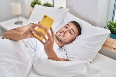 Genç İspanyol adam yatakta akıllı telefon kullanıyor.