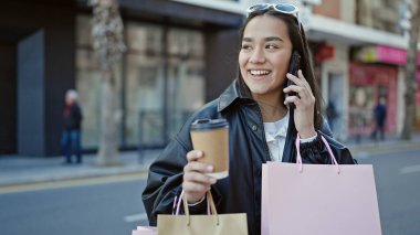 Güzel İspanyol bir kadın akıllı telefondan konuşuyor. Elinde alışveriş poşetleri ve sokakta kahve var.