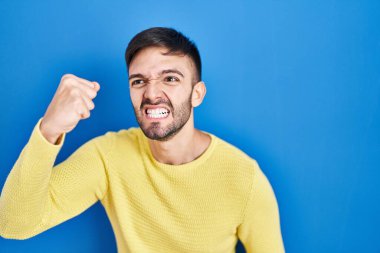 Hispanic man standing over blue background angry and mad raising fist frustrated and furious while shouting with anger. rage and aggressive concept. 