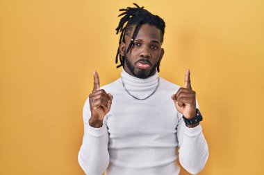 Sarı arka planda balıkçı yaka kazak giyen Afrikalı adam üzgün ve üzgün görünüyor, parmaklarıyla yön gösteriyor, mutsuz ve depresif.. 