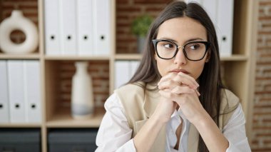 Young beautiful hispanic woman business worker sitting on table with relaxed expression at office clipart