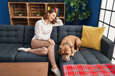 Beyaz bir kadın, evinde köpekle kanepede otururken kendinden emin bir şekilde gülümsüyor.