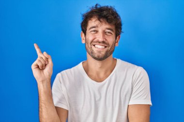 Mavi arka planda duran Latin asıllı genç adam yüzünde büyük bir gülümsemeyle, kameraya el ile işaret ederek bakıyor.. 
