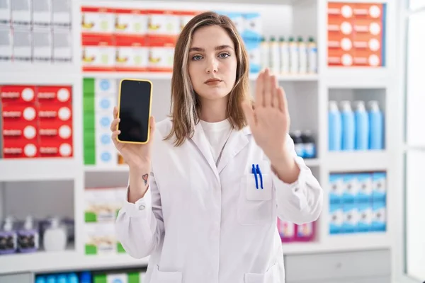 在药店工作的金发白人妇女展示智能手机屏幕时 她用张开的手做着停止手势 脸上带着严肃而自信的表情和防御姿态 — 图库照片