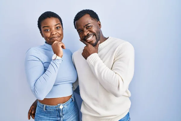 年轻的非洲裔美国夫妇手托着下巴站在蓝色的背景上思考着问题 沉思着 微笑和体贴的脸 怀疑概念 — 图库照片