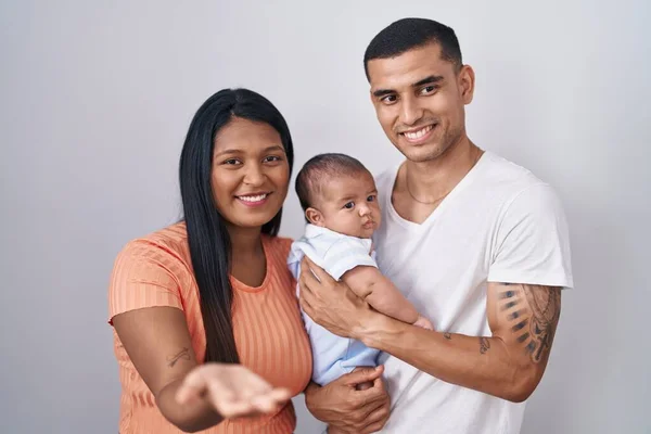 孤立した背景の上に一緒に立って赤ちゃんを持つ若いヒスパニック系のカップル笑顔陽気な提供ヤシの手援助と受け入れ — ストック写真