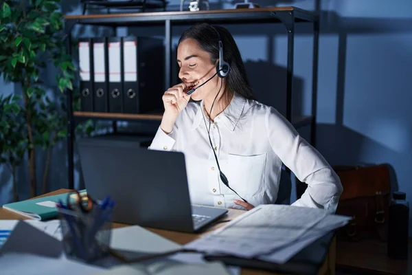 Young brunette woman wearing call center agent headset working late at night feeling unwell and coughing as symptom for cold or bronchitis. health care concept.