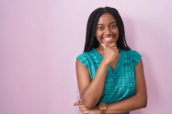 Young african american with braids standing over pink background looking confident at the camera smiling with crossed arms and hand raised on chin. thinking positive.