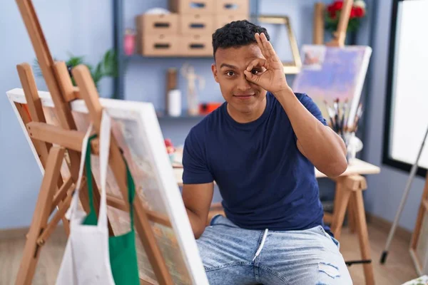 若いですヒスパニック男絵画で座っていますでアートスタジオ笑顔幸せありますOk Signとともに手で目を通して指を見て — ストック写真