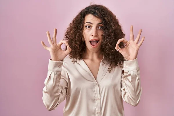 头发卷曲的西班牙裔女人站在粉红的背景上 看起来惊讶而震惊 用手指做了个认可的符号 疯狂的表达 — 图库照片