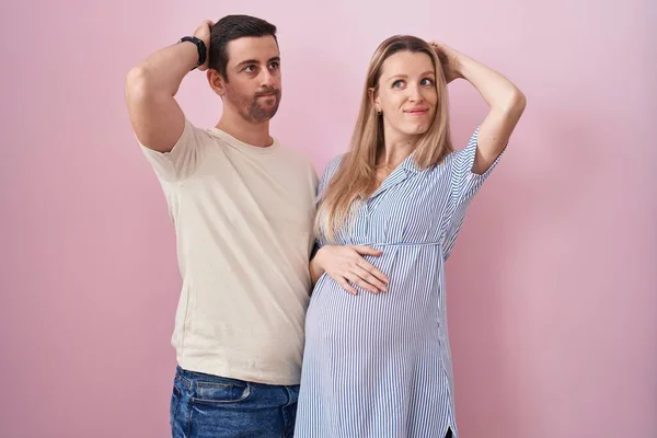 ピンク色の背景の上に立つ赤ん坊を期待している若いカップルは混乱し 疑問に思う 疑いもなく頭に手を置いて考える 包括的な概念は — ストック写真