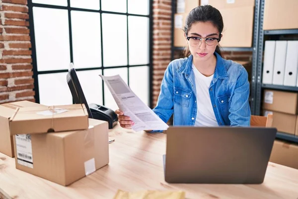Young beautiful hispanic woman ecommerce using laptop reading document at office