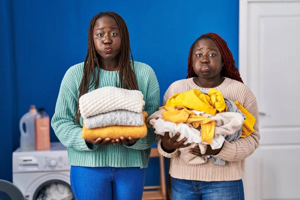 Две Африканки Держащие Сложенное Белье После Глажки Пухлых Щек Забавным — стоковое фото