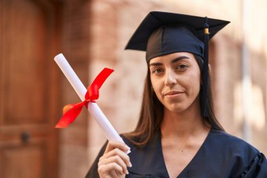 Young hispanic woman wearing graduated uniform holding diploma at university