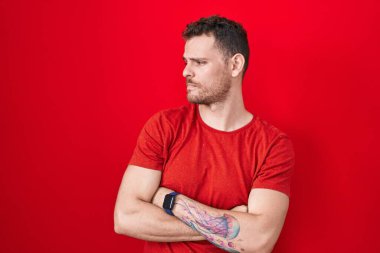 Young hispanic man standing over red background looking to the side with arms crossed convinced and confident 