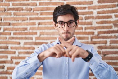 Tuğla duvar arkasında duran İspanyol genç adam. Reddedilme ifadesi. Eksi işareti yapan parmaklar. 