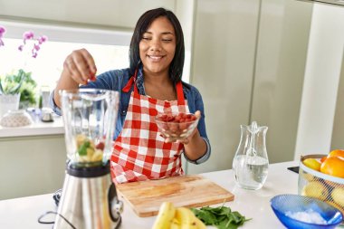 Hispanic brunette woman preparing fruit smoothie with raspberries at the kitchen