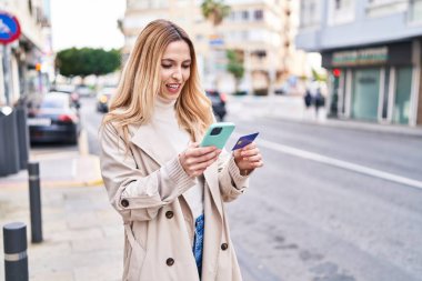 Young blonde woman using smartphone and credit card at street