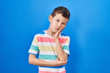 Mavi arka plan üzerinde duran genç beyaz çocuk çapraz kollarla depresyon sorunlarından sıkılmış ve yorgun görünüyor.. 