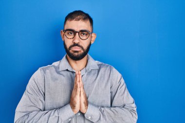 Orta Doğulu sakallı bir adam mavi arka planda duruyor. Elleriyle yalvarıyor ve dua ediyor. Yüzünde umut ifadesi var. Yalvarıyor. 