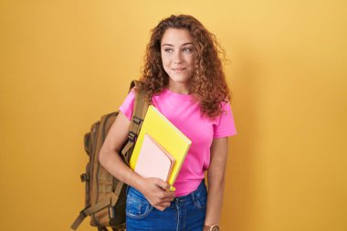 Sırt çantası takan, elinde kitap tutan, gülümseyen ve kafasını çeviren beyaz bir kadın.. 