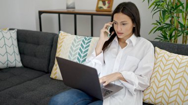 Genç, güzel, İspanyol bir kadın akıllı telefonuyla evde dizüstü bilgisayar kullanıyor.