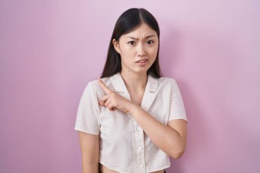 Chinese young woman standing over pink background pointing aside worried and nervous with forefinger, concerned and surprised expression 