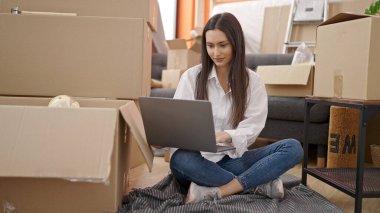 Young beautiful hispanic woman using laptop sitting on floor at new home