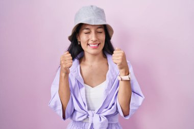 Pembe arka planda duran genç İspanyol kadın. Başarı için heyecanlı şapkalar takıyor. Kolları havada ve gözleri kapalı zaferi kutluyor. Kazanan konsept. 