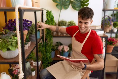 Young hispanic man florist reading book holding plant of shelving at flower shop