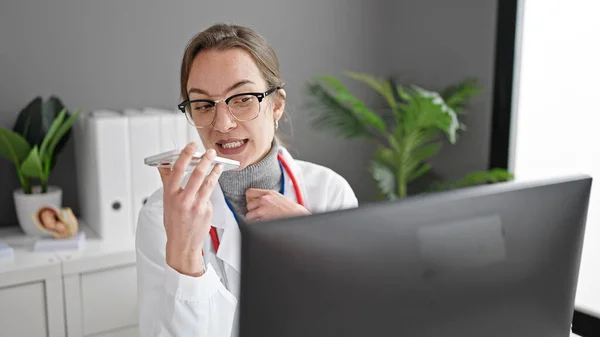 年轻的高加索女医生在诊所用电脑用智能手机传送语音讯息 — 图库照片
